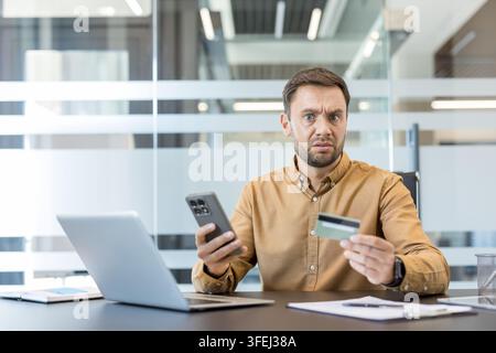 Stressed man looking at the camera with a surprised expression, holding a smartphone and credit card, experiencing online payment problems while sitting at his office desk with a laptop Stock Photo