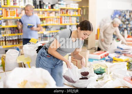 Careful young woman purchaser putting beans into plastic bag held in hand in supermarket Stock Photo