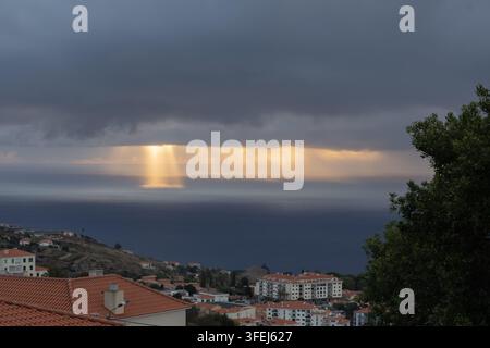 A dramatic dawn over Madeira shows golden sunlight streaming through heavy clouds and reflecting across the calm surface of the Atlantic Ocean. Stock Photo