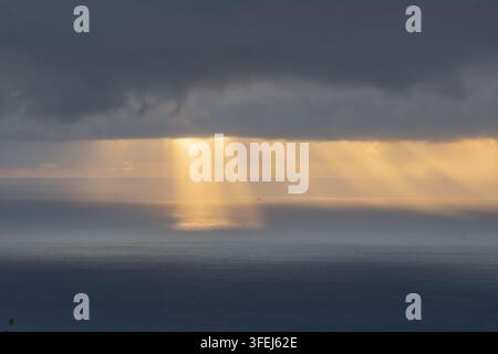 A dramatic dawn over Madeira shows golden sunlight streaming through heavy clouds and reflecting across the calm surface of the Atlantic Ocean. Stock Photo