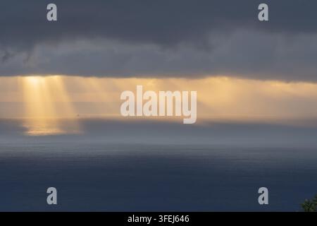 A dramatic dawn over Madeira shows golden sunlight streaming through heavy clouds and reflecting across the calm surface of the Atlantic Ocean. Stock Photo