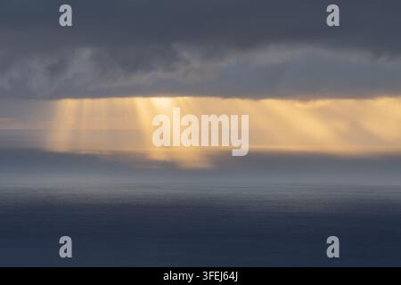 A dramatic dawn over Madeira shows golden sunlight streaming through heavy clouds and reflecting across the calm surface of the Atlantic Ocean. Stock Photo