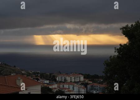 A dramatic dawn over Madeira shows golden sunlight streaming through heavy clouds and reflecting across the calm surface of the Atlantic Ocean. Stock Photo