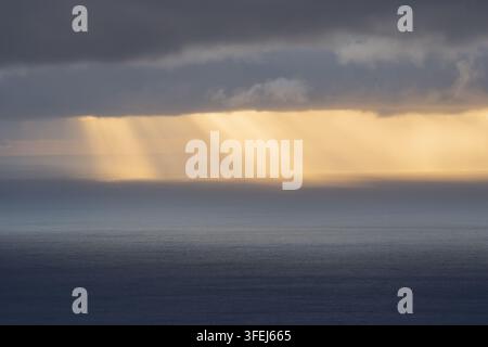 A dramatic dawn over Madeira shows golden sunlight streaming through heavy clouds and reflecting across the calm surface of the Atlantic Ocean. Stock Photo