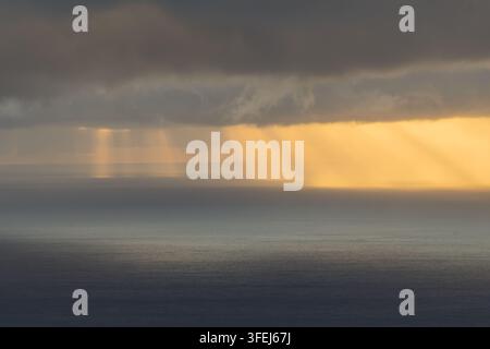 A dramatic dawn over Madeira shows golden sunlight streaming through heavy clouds and reflecting across the calm surface of the Atlantic Ocean. Stock Photo