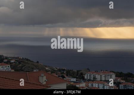 A dramatic dawn over Madeira shows golden sunlight streaming through heavy clouds and reflecting across the calm surface of the Atlantic Ocean. Stock Photo