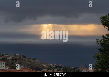 A dramatic dawn over Madeira shows golden sunlight streaming through heavy clouds and reflecting across the calm surface of the Atlantic Ocean. Stock Photo