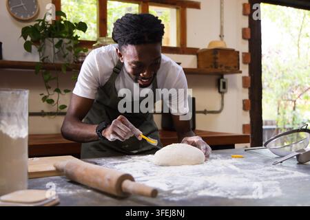 Man wearing apron scoring round loaf on wooden countertop at rustic kitchen, using small blade Stock Photo