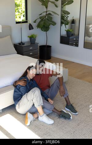 Diverse couple sitting on rug near bed in bedroom, woman leaning against man wearing denim jacket Stock Photo