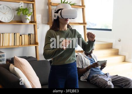 Couple at home, woman wearing white VR headset gesturing while man seated on sofa using tablet Stock Photo