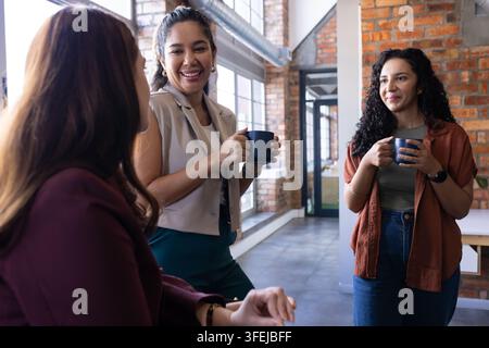 Diverse female coworkers chatting and holding coffee mugs in business-casual office with brick wall Stock Photo