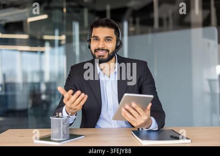 Professional man wearing a headset and suit working in an office, holding a tablet and gesturing, providing customer service and technical support with a positive expression Stock Photo