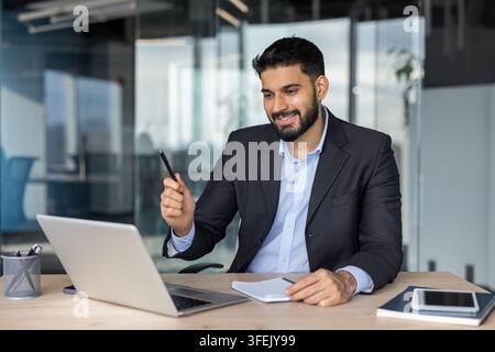 Businessman smiling and pointing with a pen during a video conference on his laptop, working on a wooden desk in a modern corporate office environment Stock Photo