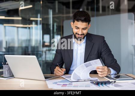 Smiling businessman reviewing financial reports and paperwork while working on laptop and tablet in a modern corporate workspace, focused on planning, strategy and productivity Stock Photo