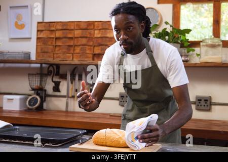 African American man in apron shaping round loaf on cutting board in bakery, holding towel. Baker, kitchen, baking, bread, wooden, warm, calm Stock Photo