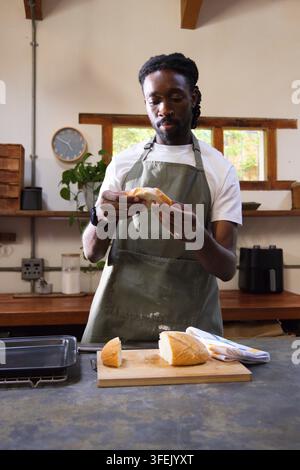 African American man in green apron inspecting partially sliced loaf on cutting board on counter Stock Photo