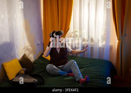 Stylish young woman uses VR glasses in a warm bedroom interior. Digital interaction and future technologies in a home setting. Mid shot. Stock Photo