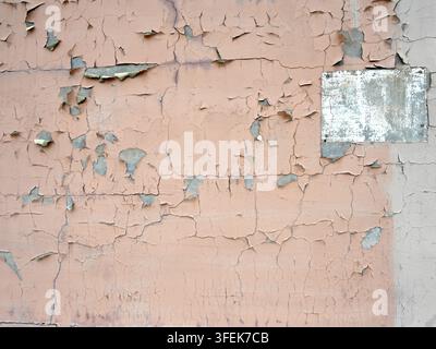 Weatherworn tan painted concrete wall in horizontal close-up with cracked peeling paint, exposed cement, chipped white painted rectangle Stock Photo
