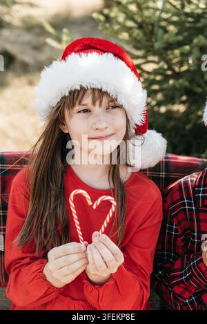 Christmas in July with smiling girl wearing Santa hat and holding heart made from candy canes in sunny summer garden beside evergreen Christmas tree. Stock Photo