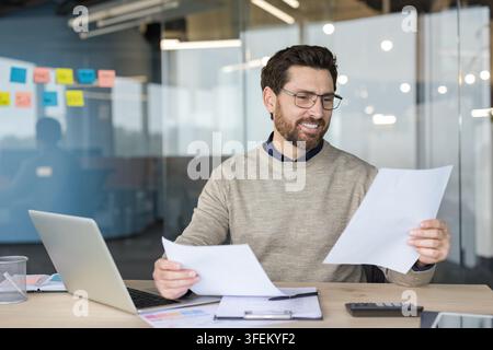 Businessman in a modern office smiling while reviewing financial reports and analyzing data on laptop and paperwork, focused on planning strategy and accounting details Stock Photo