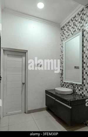 Modern minimalist bathroom interior featuring a dark vanity with white washbasin, large mirror, mosaic tile backsplash, and a closed white door. Stock Photo