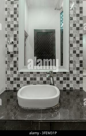 Modern bathroom interior featuring a white vessel sink on a dark marble vanity, a large mirror, and a black and white mosaic tile backsplash. Stock Photo