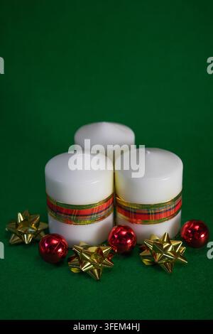 Three white candles decorated with ribbons accompanied by red baubles and gold bows on a deep green background. Concept of warmth, elegance, and Chris Stock Photo