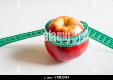 Concept of healthy lifestyle, dieting and weight loss. Fresh red apple wrapped with green centimeter tape measure on a neutral bright surface. Stock Photo