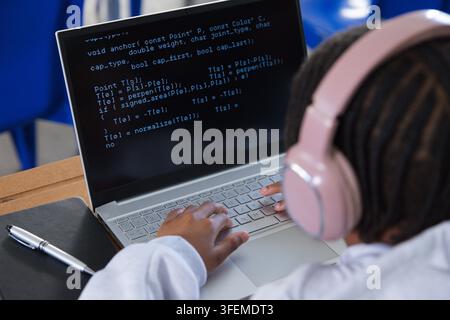 African American male youth typing code on silver laptop with pink over-ear headphones in classroom Stock Photo
