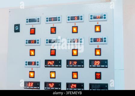 Gray metal control panel fitted with numbered rocker switches, glowing orange indicators, and red LED digital displays showing temperature values. Stock Photo