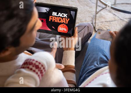 Couple browsing Black Friday 50% on tablet while holding credit card on sofa with knit sweaters Stock Photo