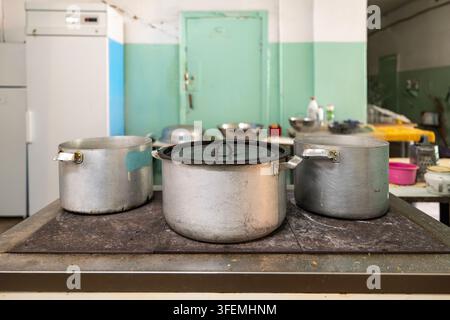 Commercial kitchen interior with large metal pots on a heavy stove and various utensils, a vintage refrigerator and a green door in the background for Stock Photo