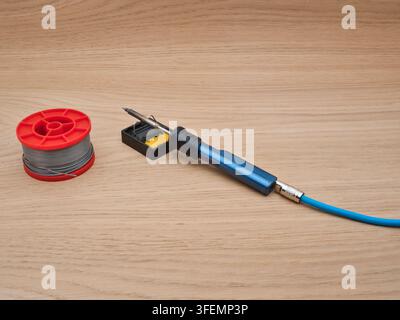 Soldering Iron, Solder, and Stand - A blue soldering iron rests on its stand with a yellow sponge, next to a red spool of solder wire, all placed on a Stock Photo