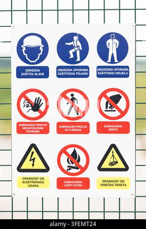 A white safety sign panel mounted on a fence displaying mandatory PPE icons, prohibition symbols and hazard warnings in blue, red and yellow to commun Stock Photo