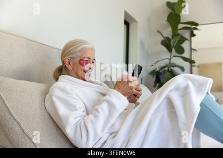 Senior woman reclining on sofa at home wearing robe and pink under-eye patch holding smartphone Stock Photo