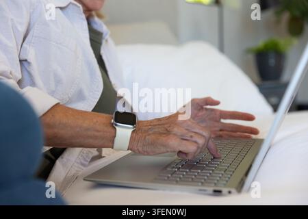 Senior woman typing on silver laptop while wearing smartwatch with white band on white bedding Stock Photo