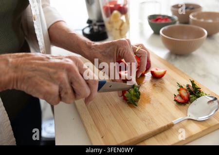 Senior woman in green top trimming, slicing strawberries on wood board in kitchen with small knife Stock Photo