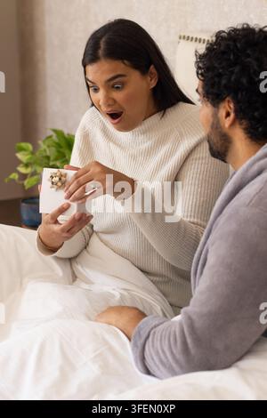 African American couple sitting on bed, woman holding gift box, left ring visible, man watching Stock Photo