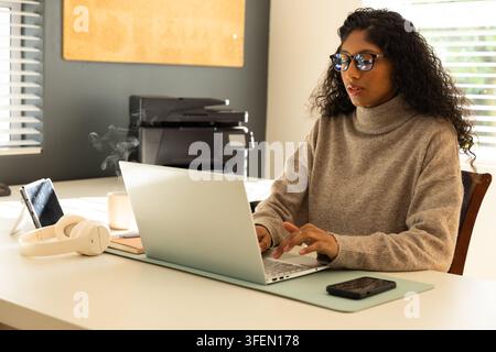 African American woman in turtleneck typing at home office on laptop with printer, copy space Stock Photo