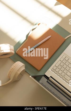 Tan leather notebook resting on green pad near window, pen lying diagonally, laptop and headphones Stock Photo