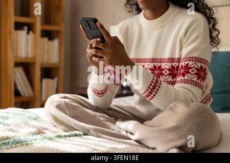 Woman sitting on bed holding smartphone and scrolling wearing knit sweater and showing gold ring Stock Photo