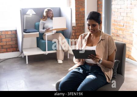 Diverse female coworkers working in lounge woman using tablet, booth worker using laptop Stock Photo