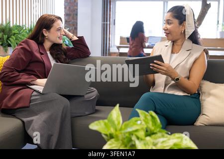 Female colleagues in blazers sitting on gray sofa in lounge, one holding tablet other using laptop Stock Photo