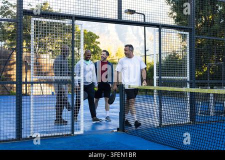 Diverse male friends entering gate onto blue court holding paddles and pickleball in shoes Stock Photo