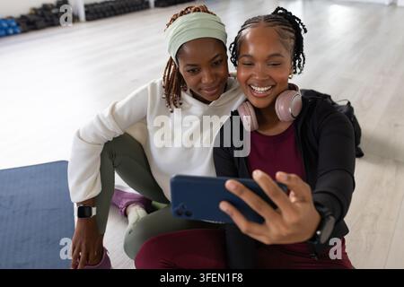 Two African women sitting on yoga mat in gym taking selfie with phone, pink headphones, dumbbells Stock Photo