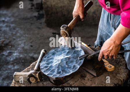 A copper smith uses a hammer and tongs to work hot copper fresh from the forge at a family owned copper workshop in Santa Clara del Cobre, Mexico. Copper-smithing has been practiced in the area since pre-Hispanic times and 82% of the towns population is involved in crafting copper items. Stock Photo