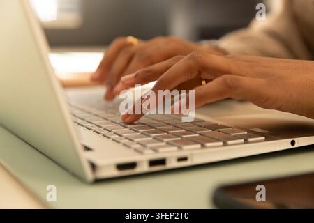 African American woman typing on laptop keyboard at desk, beige long-sleeves and gold ring visible Stock Photo