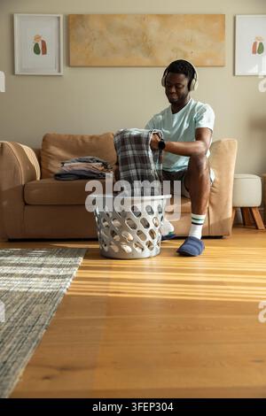 African American man folding laundry from gray basket on beige sofa with headphones, copy space. Sunlit, cozy, home, livingroom, abstract, rug, clothe Stock Photo