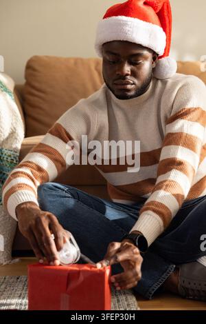 African man in Santa hat wrapping red gift box using tape dispenser before beige couch Stock Photo