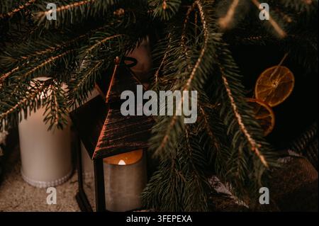 Vintage metal lantern with a burning candle standing among fresh fir branches with dried orange slices and a pillar candle. Warm cozy Christmas home d Stock Photo